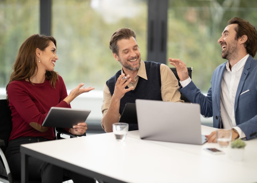 Three colleagues talking across a meeting table with a laptop open between them.